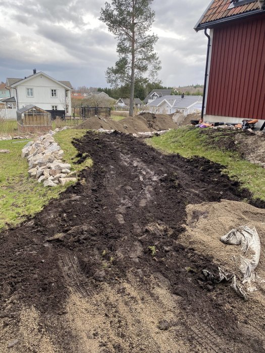 Nygrävd jord och grushög i trädgård med bakgrund av bostadshus och träd.