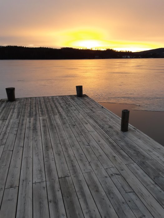 Träbrygga som leder ut till stilla vatten under en solnedgång med skog i bakgrunden.