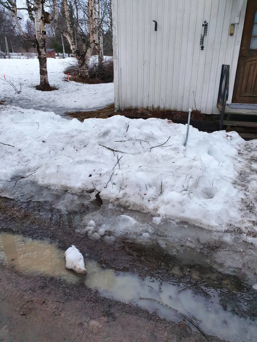 Vattenansamling vid husgrund med smältande snö och planteringar synliga.