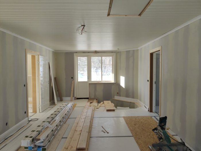 Renoveringsarbete i rum med plankor och byggmaterial på golvet och ett lågt placerat fönster på väggen.