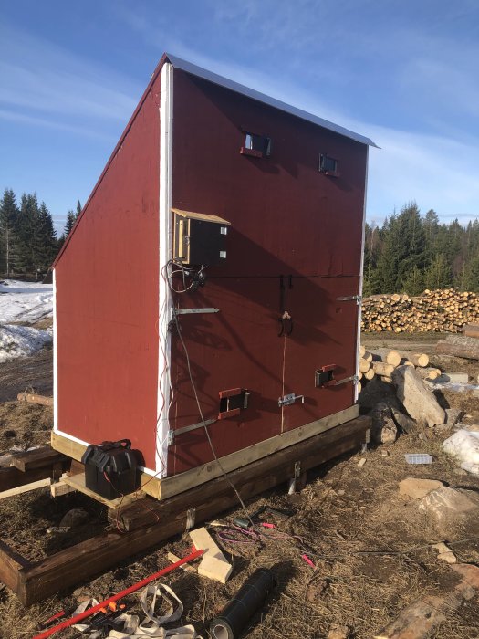 Röd byggnad på ny grund med jordankare, verktyg och byggmaterial på marken, med skog i bakgrunden.