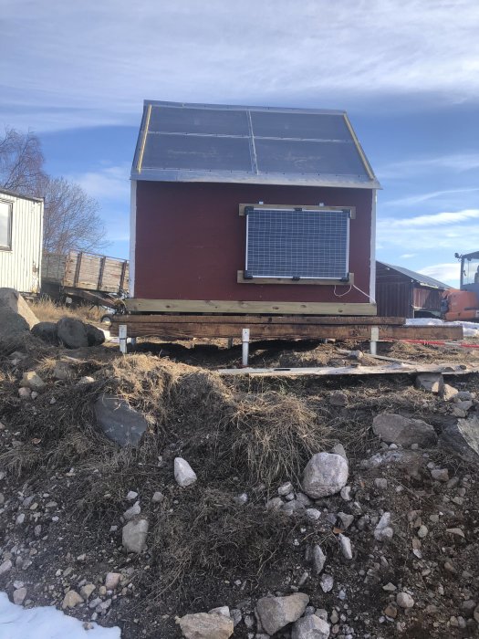 Rödfärgad byggnad upphöjd på grund av jordankare med solpanel på vägg och snötäckt mark.