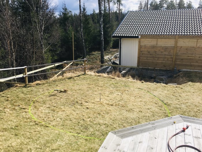 Altanprojekt med markerad cirkel på gräsmatta, staket och skog i bakgrunden, samt ett förråd till höger.