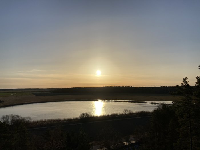 Solnedgång över ett lugnt vattendrag med reflektion av solen och omgivande skog och fält.