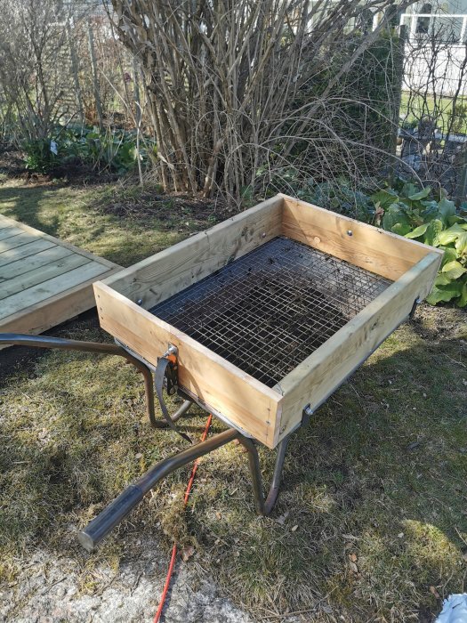 Hemmagjort såll på en skottkärra gjort av träram och nät, omgivet av gräs och buskar.