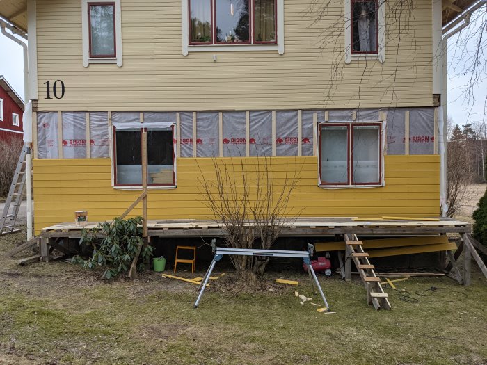 Delvis monterad gul träpanel på hus exteriör med byggmaterial och verktyg framför.