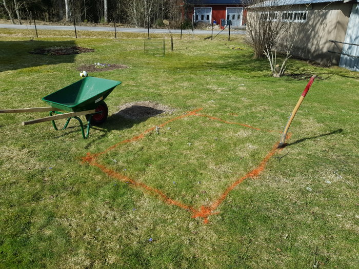 Uppmärkt gräns med orange sprayfärg för sandlåda nära ett hus, med skottkärra och spade på gräsmattan.