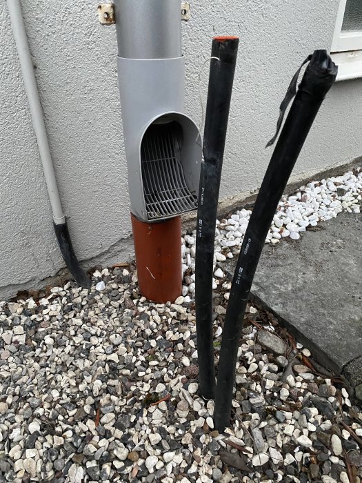 Två svarta elkablar sticker ut ur marken vid en husvägg bredvid en grå nedrör.