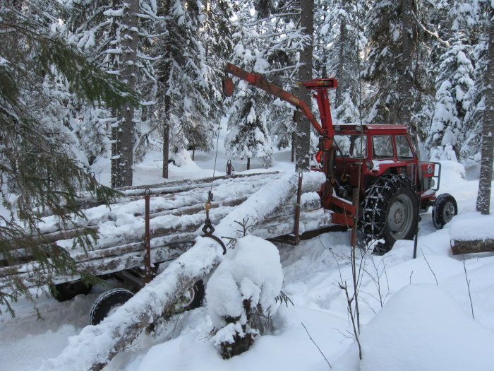 Gammal traktor med vajerkran och släpvagn lastad med sågstockar i snötäckt skogsmiljö.