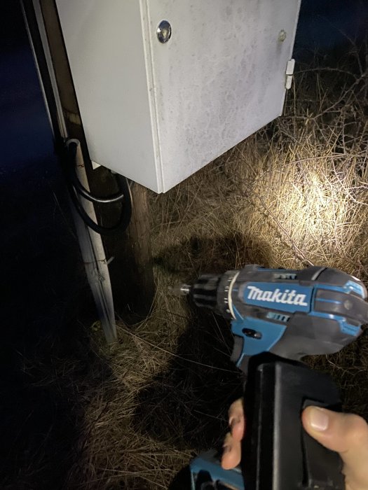 En Makita skruvdragare hålls framför en elskåp utomhus på natten.