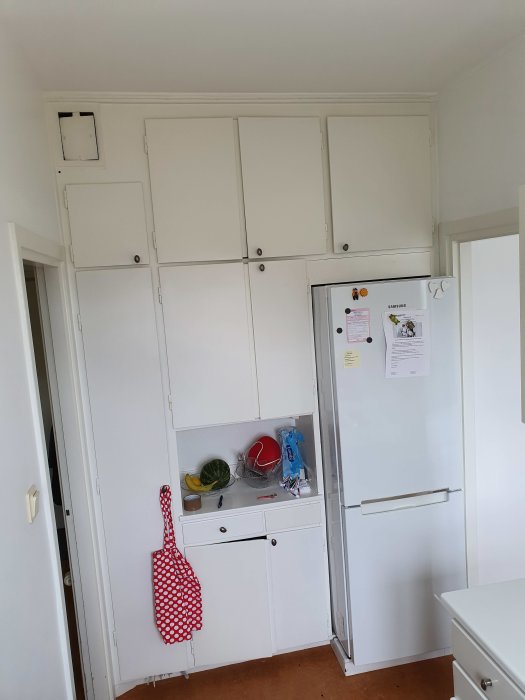 Vitt kök med platsbyggda skåp och kylskåp, i behov av renovering och anpassning till takhöjd.