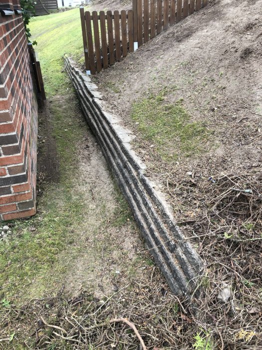 Gammal låg stödmur längs en slänt med nytt staket ovanför, behöver renovering.