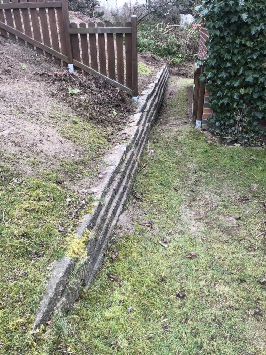 Gammal U-formad stödmur av sten i en trädgård med en slänt och staket i bakgrunden.