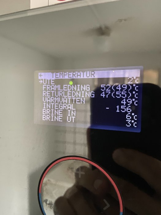 Display av värmeväxlare som visar olika temperaturer och värden, reflektion av personens hand.