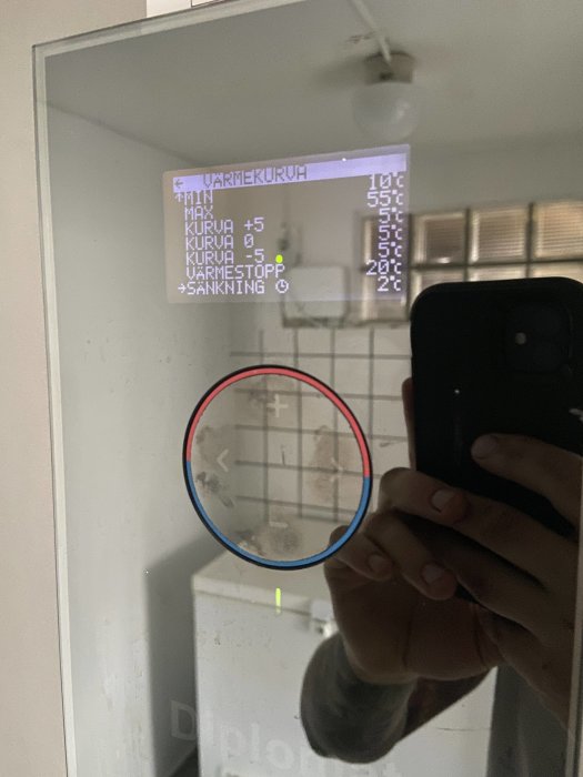 Person som håller en mobiltelefon och reflexion av en upplyst termostatdisplay i ett fönster.