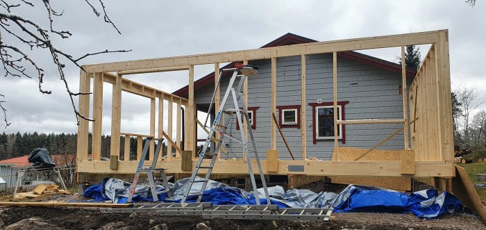 Pågående bygge av en yttervägg i träkonstruktion med existerande grå hus i bakgrunden, verktyg och en stege syns.