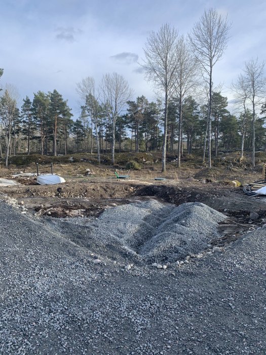 Tomt med berg och grus där arbete påbörjats för byggprojekt, omgivet av skog och blå himmel.