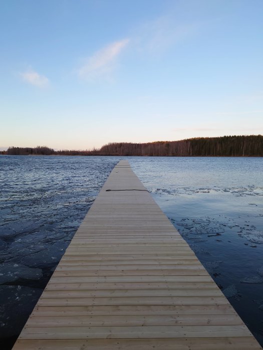 Nybyggd träbrygga som sträcker sig ut över en delvis isbelagd sjö med skog i bakgrunden.