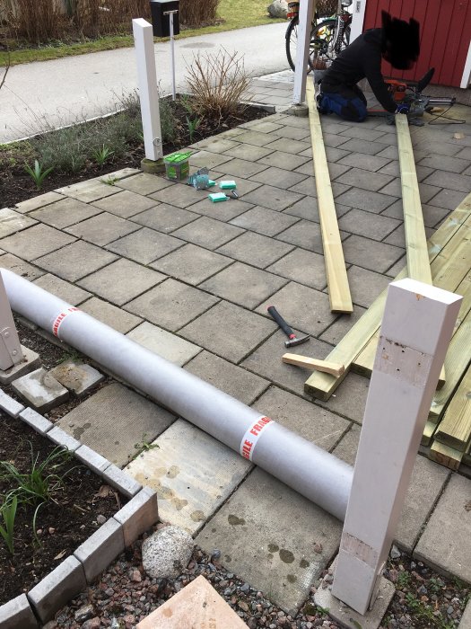 Person som arbetar med att såga träplankor på en uppfart med stenplattor, byggmaterial och verktyg syns.