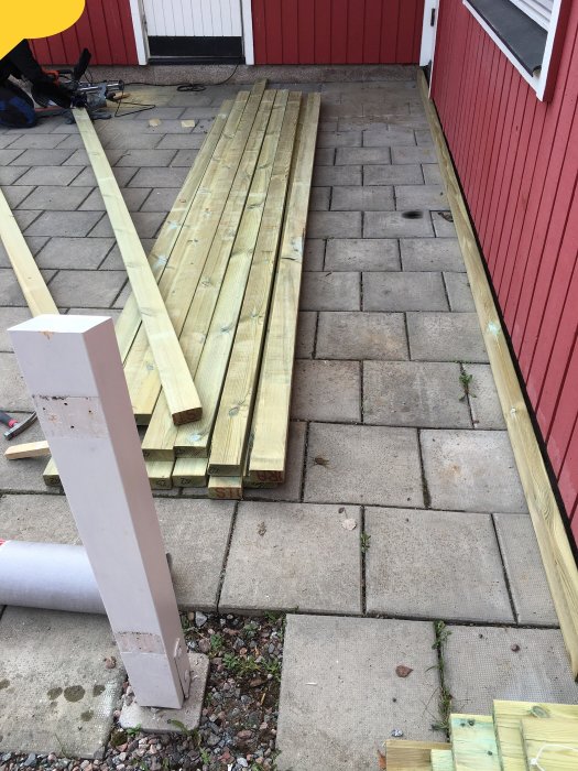 Trämaterial staplat på stenläggning vid röd husvägg, byggprojektets första dag.