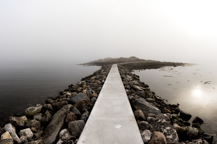 Betonstig som sträcker sig över en stenig strand och in i dimman över vattnet.