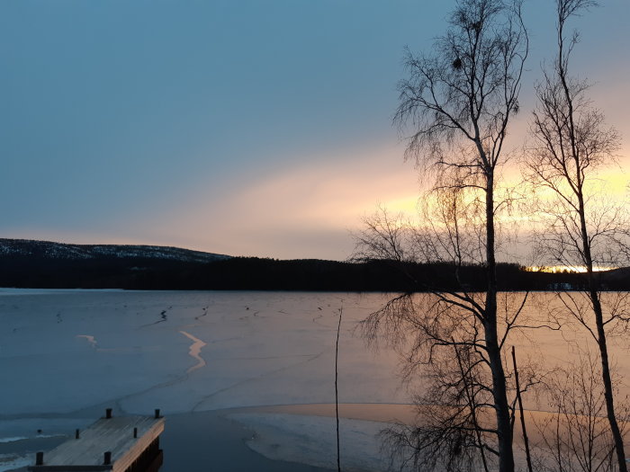 Solnedgång över en frusen sjö med siluetter av träd och ett bryggans hörn i förgrunden.