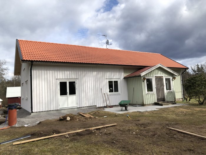 Renoverat hus med ny panel och röd takbeklädnad, byggmaterial och skottkärra utanför.