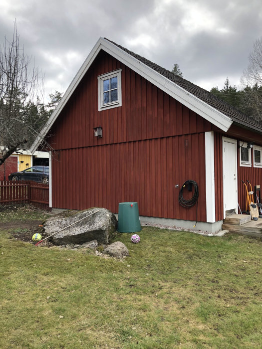 Röd träbyggnad med vit dörr och fönster ovanför en stor sten, plats för planerad utvändig trappa.