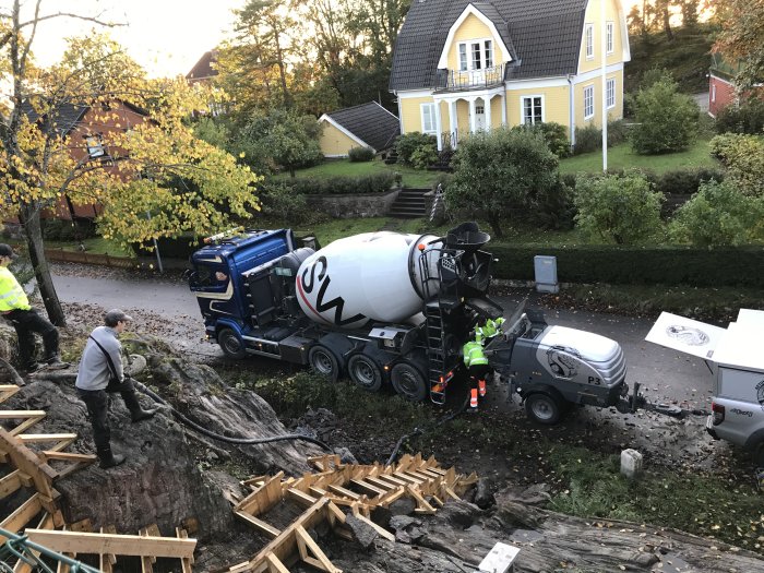 Bild på betongblandare och arbetare i ett byggprojekt vid en trappa under konstruktion, med ett hus i bakgrunden.