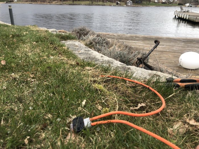 En orange slinga på grön gräsmatta med sjö och brygga i bakgrunden, motorsåg och verktyg synliga.