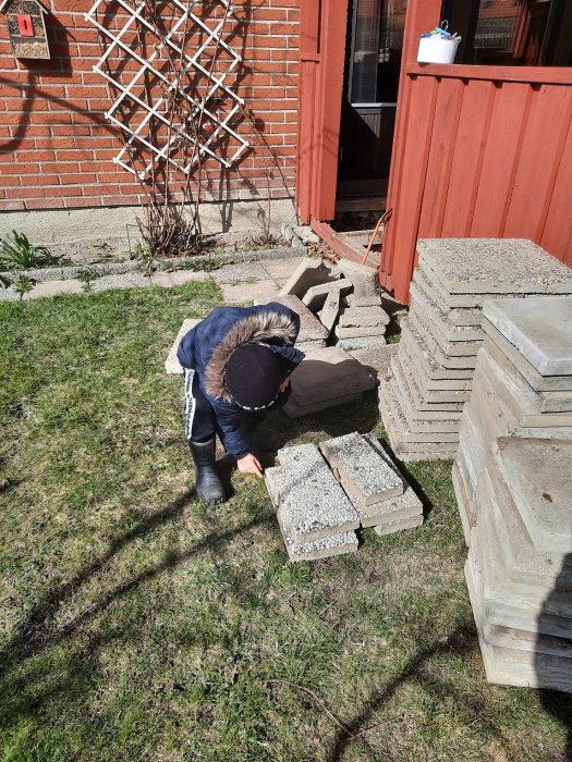 Barn hjälper till att bära trottoarstenar i trädgården, bredvid en stapel av plattor.