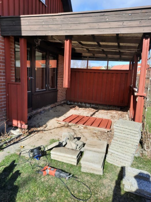 Byggplats med avlägsnade tunga betongstenar och verktyg framför ett rödfärgat hus under rivningsarbete.