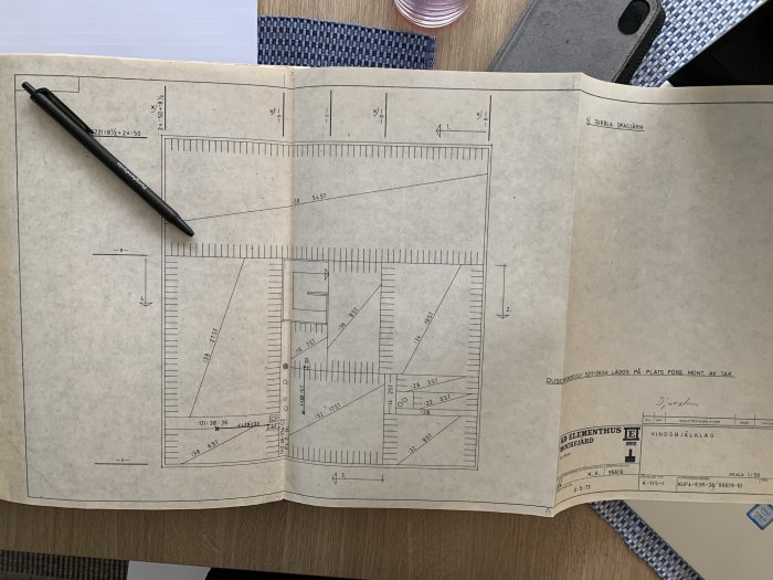 Ritning öppnad på ett bord med måttangivelser, avser planlösning av ett 1,5-planshus från AB Elementhus Mockfjärd, inklusive en penna.