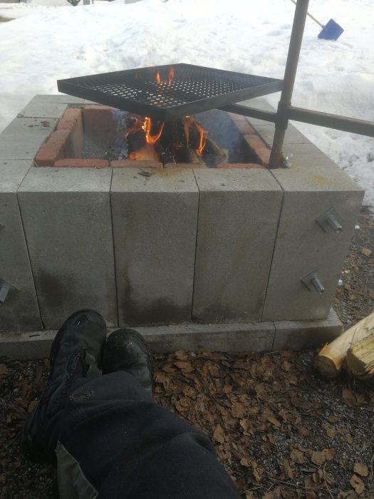 En egenbyggd uteplats med öppen eldstad av betongblock och metallgaller, med snö i bakgrunden och en person i förgrunden.