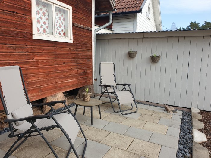 Uteplats med stolar och ett litet bord på stenplattor, två korgar med växter på en vit vägg och en rödmålad stuga till vänster.