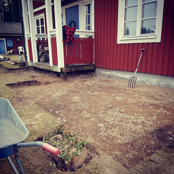 Röd stuga med förarbete för markstensläggning framför farstukvist och trädgårdsredskap.