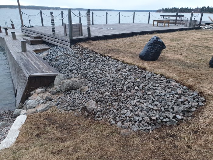 Rensad stenkant vid vatten med gräs, mossa och stor svart säck i bakgrunden.