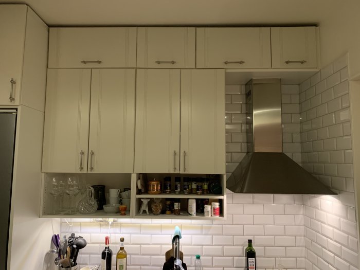 Kök med vita kakelväggar och skåp, de två nedersta skåpen saknar luckor, rörigt innehåll synligt.
