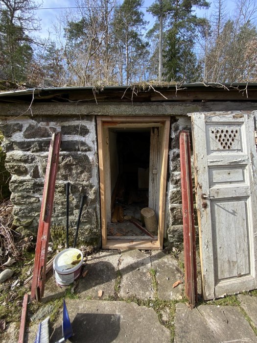 Öppen dörrkarm i en stenbyggnad, med en gammal stängd dörr till höger och renoveringsmaterial runtomkring.