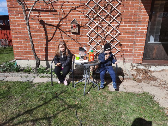 Två barn sitter vid ett trädgårdsbord framför en tegelvägg och äter korv.