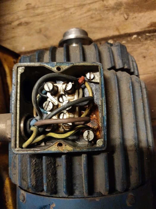 Öppen kopplingsdosa på en gammal trefasmotor med synliga kablar och anslutningar, vissa är lösa och rostiga.
