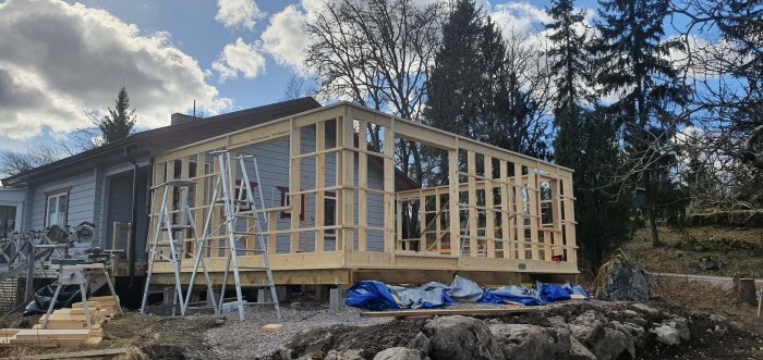 Nyreglad träkonstruktion för påbyggnad utanför ett hus, omgivet av byggmaterial och verktyg, under en delvis molnig himmel.