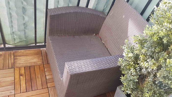 En flätad balkongsoffa i grått utan kuddar, bredvid en krukväxt på en trätrall, med balkongskydd i bakgrunden.