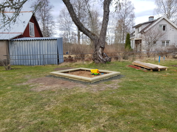 Nybyggd sandlåda med en leksakstruck i ett trädgårdsprojekt, grovsågade plankor staplade på marken.