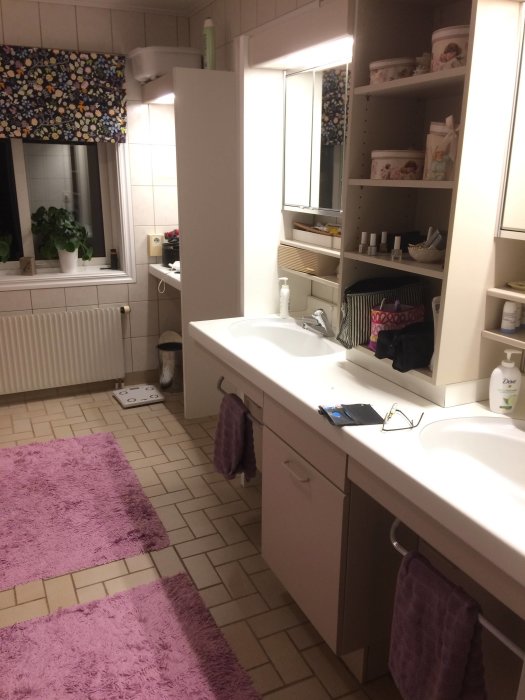 Badrum interiör med dubbel handfat, vit skåpinredning och lila badrumsmattor.