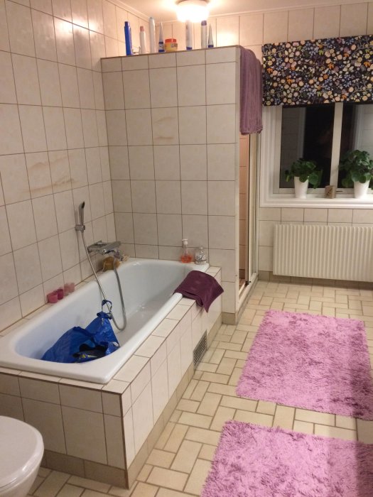 Ett badrum med badkar, kaklade väggar, två lila mattor och fönster med växter.