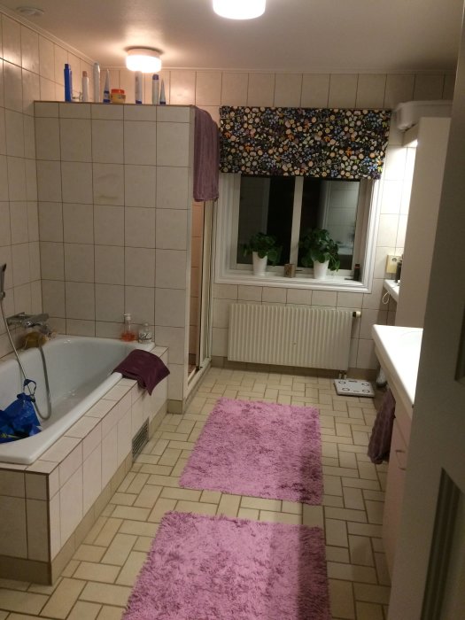 Badrum med beige kakel, badkar, två rosa badrumsmattor och fönster med blommigt draperi.