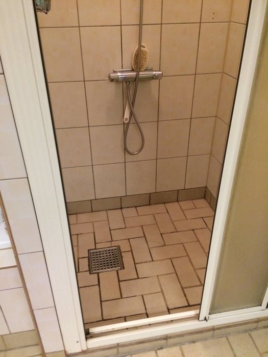 Duschhörna med beige kakel och brun mosaikgolv, handdusch och tvättborste, metallisk avloppsränna.