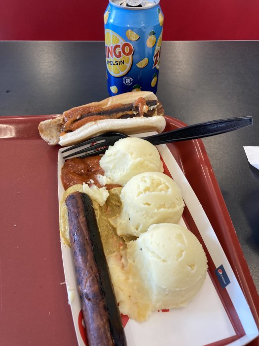 Två brända korvar med bröd, pulvermos, ketchup, senap och en burk läsk på en röd bricka.