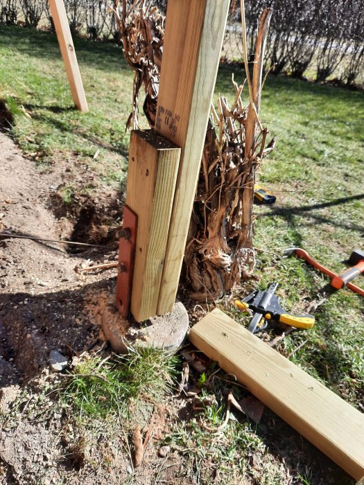 Ny trästolpe monterad vid plint och bultar med röten gammal stolpe bredvid, verktyg på gräs.